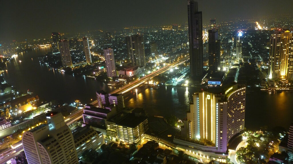 Night skyline of Bangkok Thailand with skyscrapers and the Chao Phraya River illuminated by city lights.