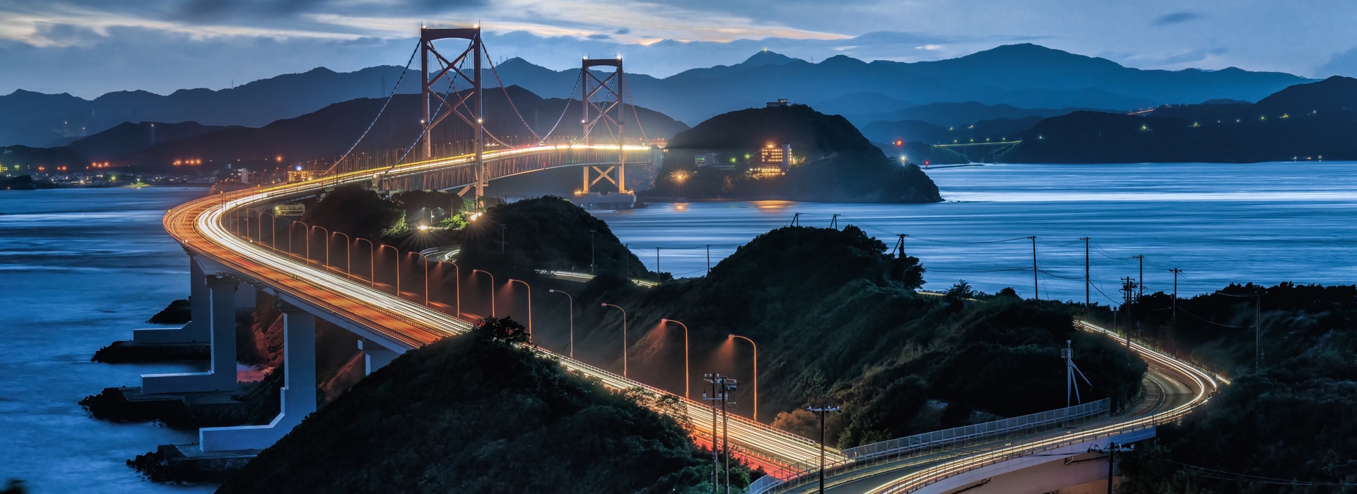 Kurushima Kaikyo Bridge illuminated at night in Japan with ocean and mountains.